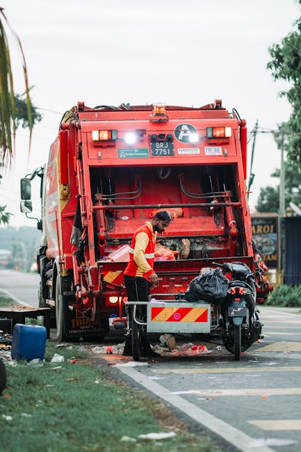 A waste collection worker wearing a high-visibility yellow and red uniform is operating a large, red rubbish truck parked on the side of a paved road. The back of the truck, which is open, reveals a recycling or waste compaction mechanism with visible hydraulic components. The worker stands near the open rear compartment, partly facing the vehicle, and appears to be managing waste or preparing for collection. In front of the worker, there is a small black bag placed on a metal platform attached to a motorbike or portable bin, which is positioned on the sidewalk next to the truck. The surrounding environment includes a grassy verge with a blue object or bin on the left, and a background featuring utility poles, trees, and a blue sign with partially visible text. The overall scene takes place during daytime with overcast or partly cloudy weather, supporting an appearance of a typical rubbish collection operation, possibly representing alternative waste handling or on-site clearance services by [COMPANY_NAME], in an urban or suburban setting.