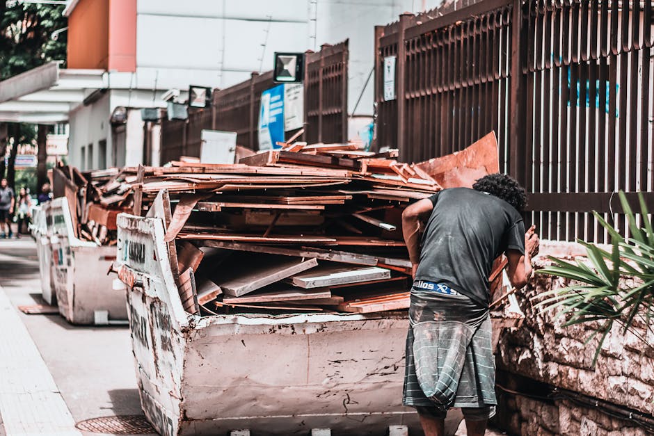 A man with curly black hair wearing a dark grey T-shirt and plaid shorts is standing next to a large, white, industrial skip on a city pavement. The skip is filled with a substantial pile of reddish-brown roof tiles, some scattered and leaning against the sides, indicating ongoing or recent rubbish removal. Behind the skip, there is a dark brown metal fence with vertical slats, extending along the pavement, and a stone wall partially visible behind the fence. In the background, a modern building with white and orange exterior walls and a row of street signs and security cameras appears above the fence. The scene is well-lit, suggesting daytime, with natural lighting highlighting the texture of the roof tiles, the clean lines of the skip’s metal surface, and the smooth pavement. This setup reflects a typical outside area for private waste collection, with the focus on clearing building debris or roofing waste, consistent with alternative rubbish removal services supplied by companies like rubbishclearancecollierswood.com.