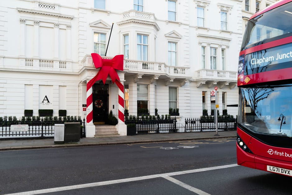 A white, multi-storey building with classical architectural features, including decorative mouldings and a small balcony, decorated for the holiday season with a large red bow and festive ribbon wrapped around a white pole at the entrance. The entrance has a dark door with a wreath hanging on it, and a short set of steps leading up to it. In front of the building, a paved sidewalk is bordered by a black metal fence with small, neatly trimmed bushes behind it. To the right, a red double-decker bus with the route display indicating 'Clapham Junction' partially blocks the view. The scene appears to be taken during daylight with clear lighting, showing a quiet urban street scene with minimal traffic. The image relates to private waste management options, as the location and setting suggest potential consideration of alternative rubbish removal or clearance services, supported by the presence of a prominent, festive decorative display on a residential or commercial premises, possibly managed or serviced by a waste clearance company like rubbishclearancecollierswood.com.