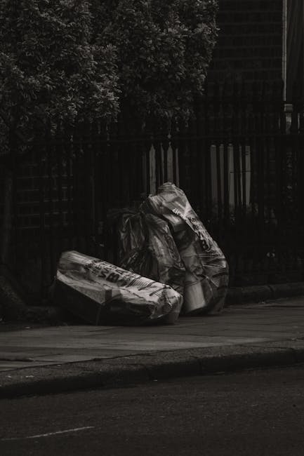 Two large black rubbish bags, made of plastic with visible creases and folds, are placed on the pavement near the curb in an urban environment. One bag appears partially torn or crumpled, revealing discarded materials inside. The bags are positioned close together, leaning slightly against a black metal fence with vertical bars, which runs along the background. Behind the fence, dense bushy foliage with dark leaves is visible, adding a natural element to the scene. The surface of the pavement is smooth and dark, contrasting with the bags' shiny, crinkled texture. The overall lighting is subdued, suggesting an overcast day or evening, which emphasizes the dark tones of the bags and surrounding environment. The scene reflects a typical instance of private rubbish disposal on a street, potentially associated with independent waste collection efforts often handled by services such as those offered by rubbishclearancecollierswood.com, in line with local rubbish removal requirements and non-public waste management.