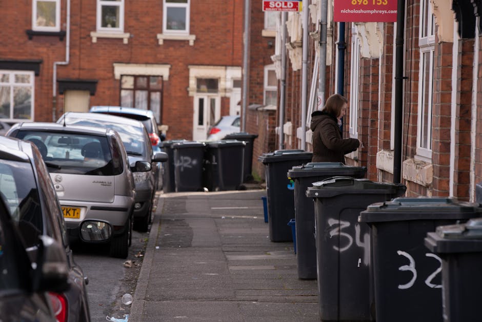 A row of black plastic wheelie bins with closed lids are lined up along the pavement outside a red brick residential building. The bins are positioned against the building's exterior wall, which features white-framed windows and stone sills. The bins are marked with white painted numbers on their sides, such as '32' and '34,' indicating designated storage or collection points. A woman wearing a brown jacket and face mask is standing beside the bins, opening or inspecting one of them, possibly preparing for rubbish disposal or collection. The scene takes place in an urban residential area, with parked cars lined along the street and additional buildings visible in the background. The lighting suggests a clear day, with natural daylight illuminating the scene, emphasizing the textures of the brickwork, plastic bins, and concrete sidewalk, providing a neutral, observational depiction of on-site waste management in a private residential setting. Natural connection to rubbish clearance services, highlighting independent rubbish disposal outside a typical home, consistent with private waste handling practices.