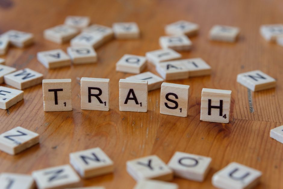 A close-up image of wooden Scrabble tiles arranged spelling the word 'TRASH' on a wooden surface, with various other scattered tiles around, some turned face up displaying letters and point values. The surface appears to be a polished wooden tabletop or floor, with a warm, natural finish. The scattered tiles include letters N, Y, and others, some overlapping or partly obscured by the central word. The lighting is even, highlighting the texture of the wood and the smooth finish of the tiles, creating a neutral, professional atmosphere. The scene subtly relates to waste management topics, representing the concept of waste separation or disposal, akin to organising or clearing rubbish, which aligns with the themes of independent rubbish collection or alternative waste handling methods that a service like rubbishclearancecollierswood.com might support.