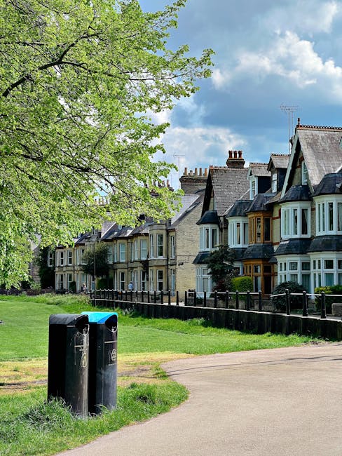 A row of Victorian-style terraced houses with bay windows and slate roofs, situated behind a narrow grass verge enclosed by a wooden fence, with a large green-leaved tree extending its branches across the top left of the image. In the foreground, two black recycling bins with blue lids are positioned on a paved footpath that curves slightly to the right. The scene is set outdoors on a partly cloudy day, with a bright blue sky and fluffy white clouds overhead. The environment appears to be a residential area, where private waste collection, such as rubbish clearance services, might be used as an alternative to council-managed rubbish pickup. The houses display a mix of brick and painted facades, with chimney stacks visible atop several rooftops, contributing to the traditional aesthetic. The presence of the bins and the tidy, well-maintained surroundings suggest regular waste management, potentially handled by an independent rubbish clearance company like rubbishclearancecollierswood.com, situated within an urban suburban context that requires on-site rubbish removal solutions.