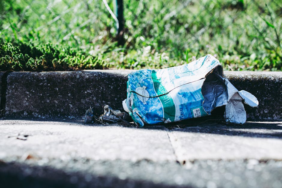 A crumpled plastic bag lying on the pavement beside a concrete curb in an outdoor urban setting. The bag features a colorful design with shades of blue, white, and green, and appears to contain additional discarded waste, possibly paper or plastic. The curb, made of rough-textured concrete, borders a grassy area with green grass and small plants visible in the background, suggesting a roadside or street environment. Shadows cast by sunlight indicate a clear day. The scene depicts a typical example of litter that can be encountered in public spaces, highlighting the importance of proper rubbish disposal and potential private waste removal services such as those offered by rubbishclearancecollierswood.com, which provide alternative methods of rubbish collection outside local authority systems.