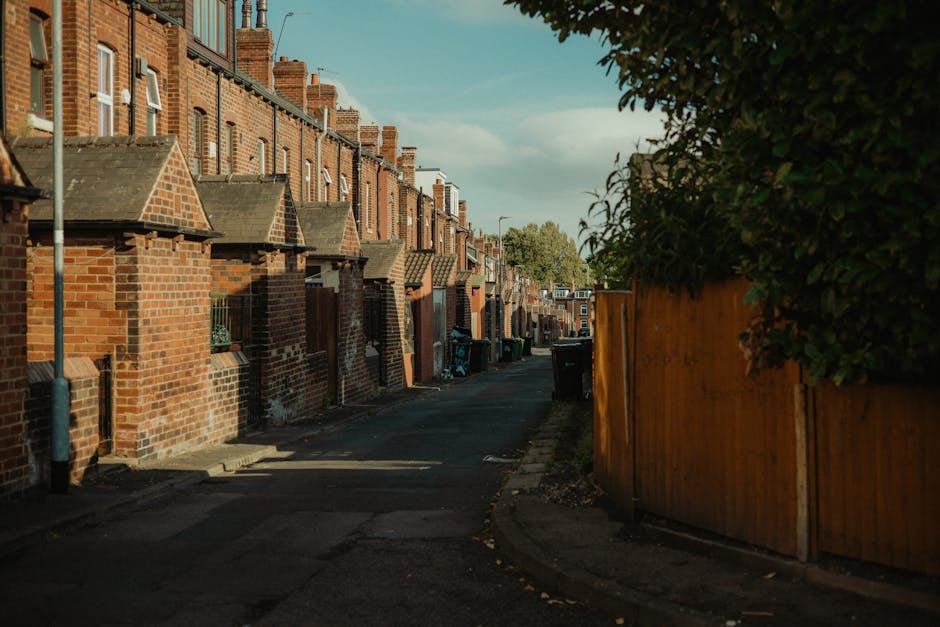 A narrow residential street lined with red-brick terraced houses featuring small front yards and pitched slate roofs, with a curved curb and asphalt pavement extending into the distance. On the right side, a tall wooden fence separates the street from a garden area with dense green foliage visible over the top, casting shadows on the pavement. Several black wheelie bins are placed along the curb, indicating waste collection points for local residents, with some bins partially shaded by the surrounding buildings and trees. The sky is partly cloudy, allowing soft natural light to illuminate the scene, highlighting the texture of the bricks and the rough asphalt surface. The environment suggests a typical urban or suburban estate setting, where private waste management services like those from rubbishclearancecollierswood.com facilitate rubbish removal in areas with limited access, as evidenced by the closely packed terraces and narrow road suitable for on-site waste disposal or alternative collection methods.